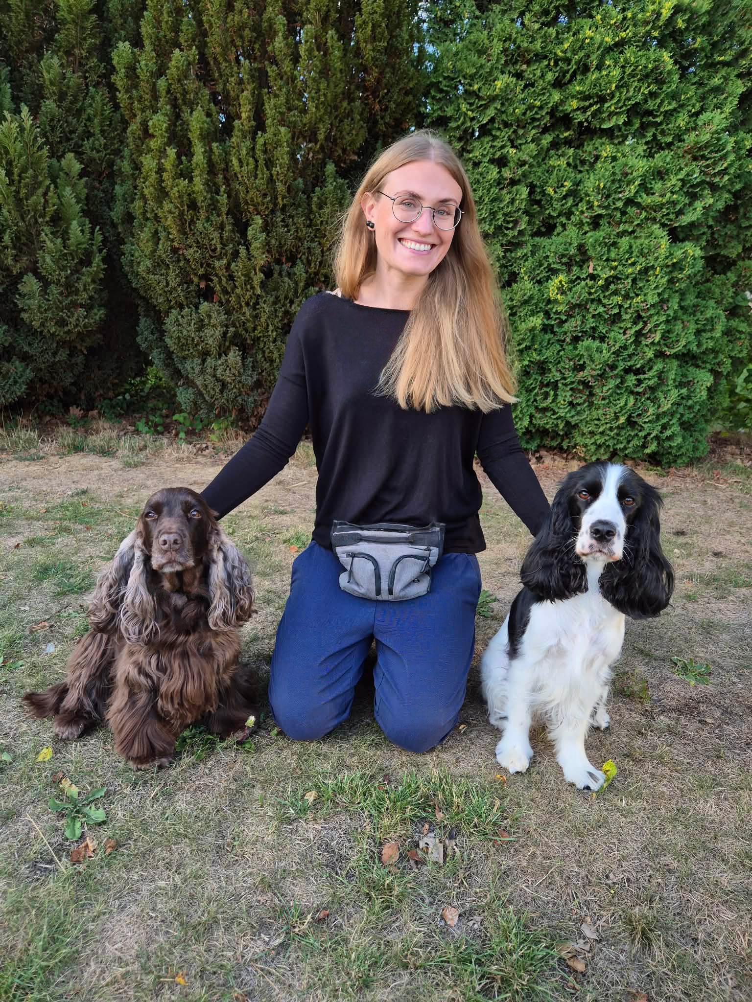 Porträttbild av en människa med långt hår, glasögon och en hundträningsväska och två hundar av rasen cocker spaniel i en trädgård.